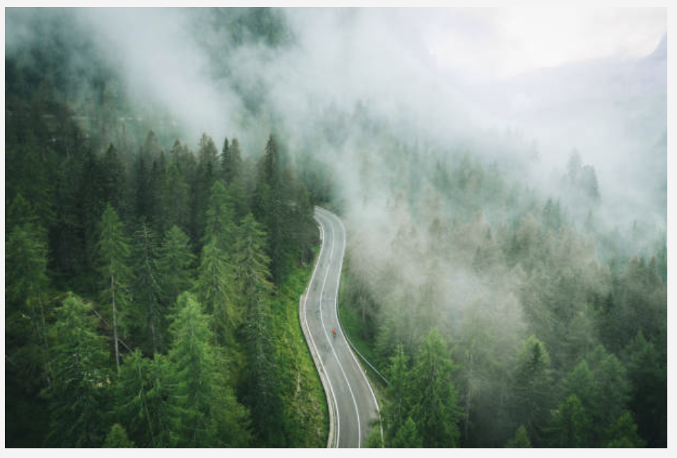The view of a winding road from above, including fog and forest and lone bicyclist.