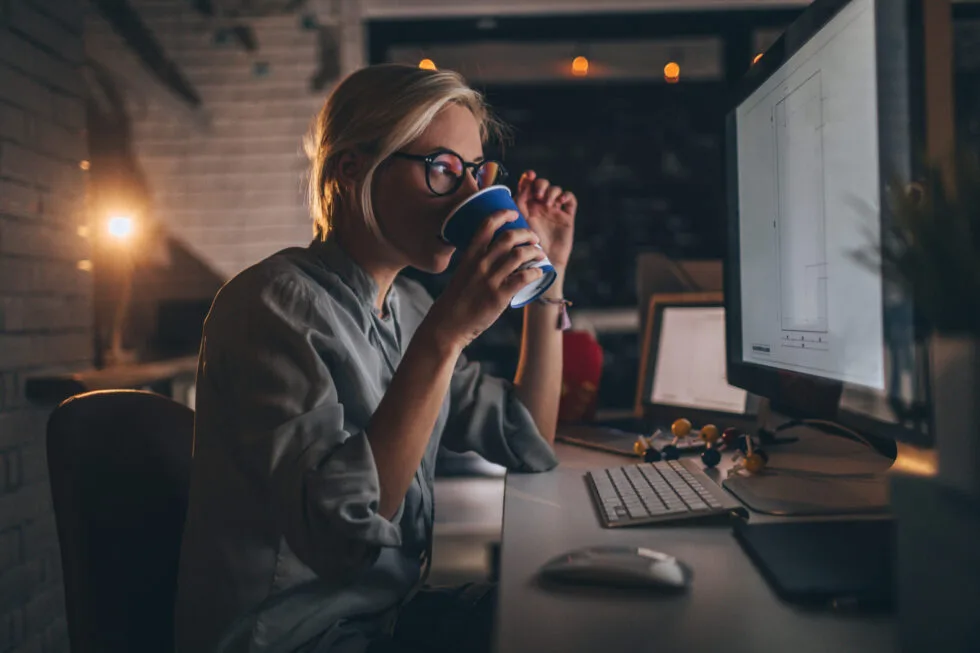 Woman at desk drinking from cup while looking at her computer monitor.