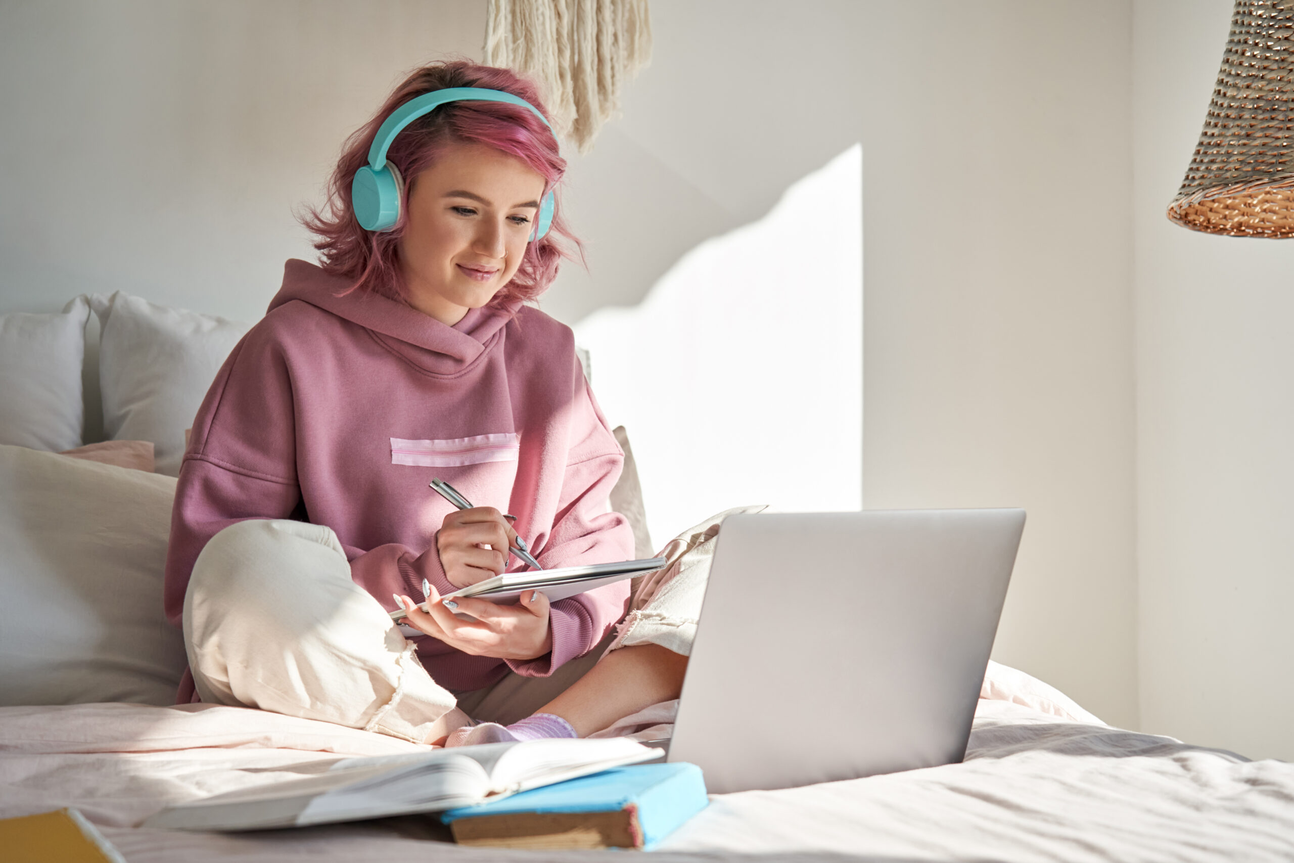 Hipster young woman sitting on her bed with headphones, taking notes on a notepad while looking at a computer