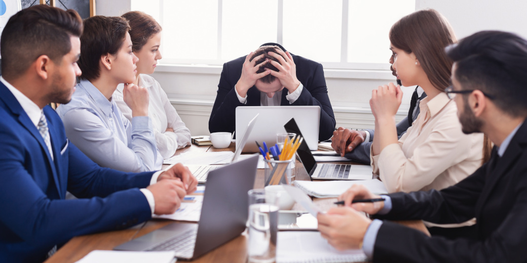 many people around a boardroom table with the person at the head of the table having his head in his hands