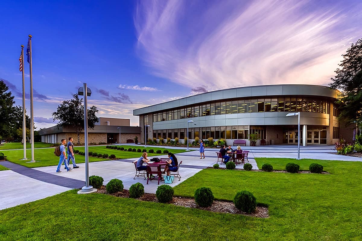 Students walking and studying outside the main building at Fox Valley Technical College’s Appleton campus during sunset. The scene features a modern curved glass building, neatly landscaped lawns, American and Wisconsin flags, and outdoor seating areas with students engaged in conversation and schoolwork.