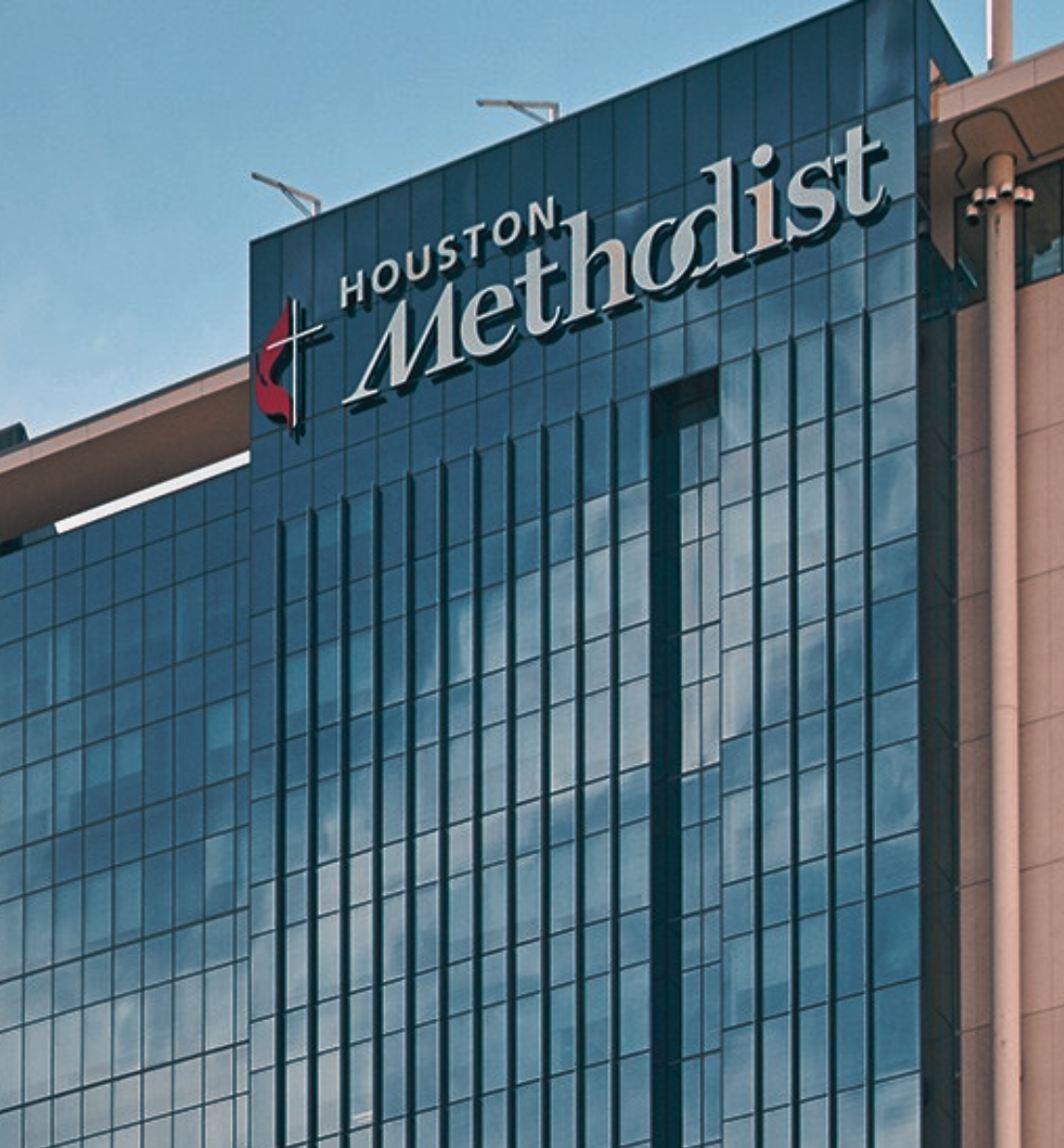 Exterior view of Houston Methodist Hospital, featuring a modern glass high-rise building with the Houston Methodist logo and cross emblem displayed near the top. The blue-tinted windows reflect the sky, and a nearby building with mirrored glass and green tree branches is partially visible on the left side.