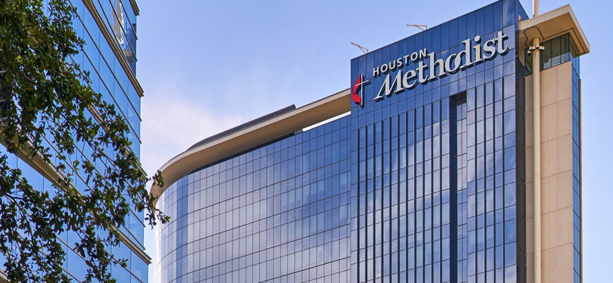 Exterior view of Houston Methodist Hospital, featuring a modern glass high-rise building with the Houston Methodist logo and cross emblem displayed near the top. The blue-tinted windows reflect the sky, and a nearby building with mirrored glass and green tree branches is partially visible on the left side.