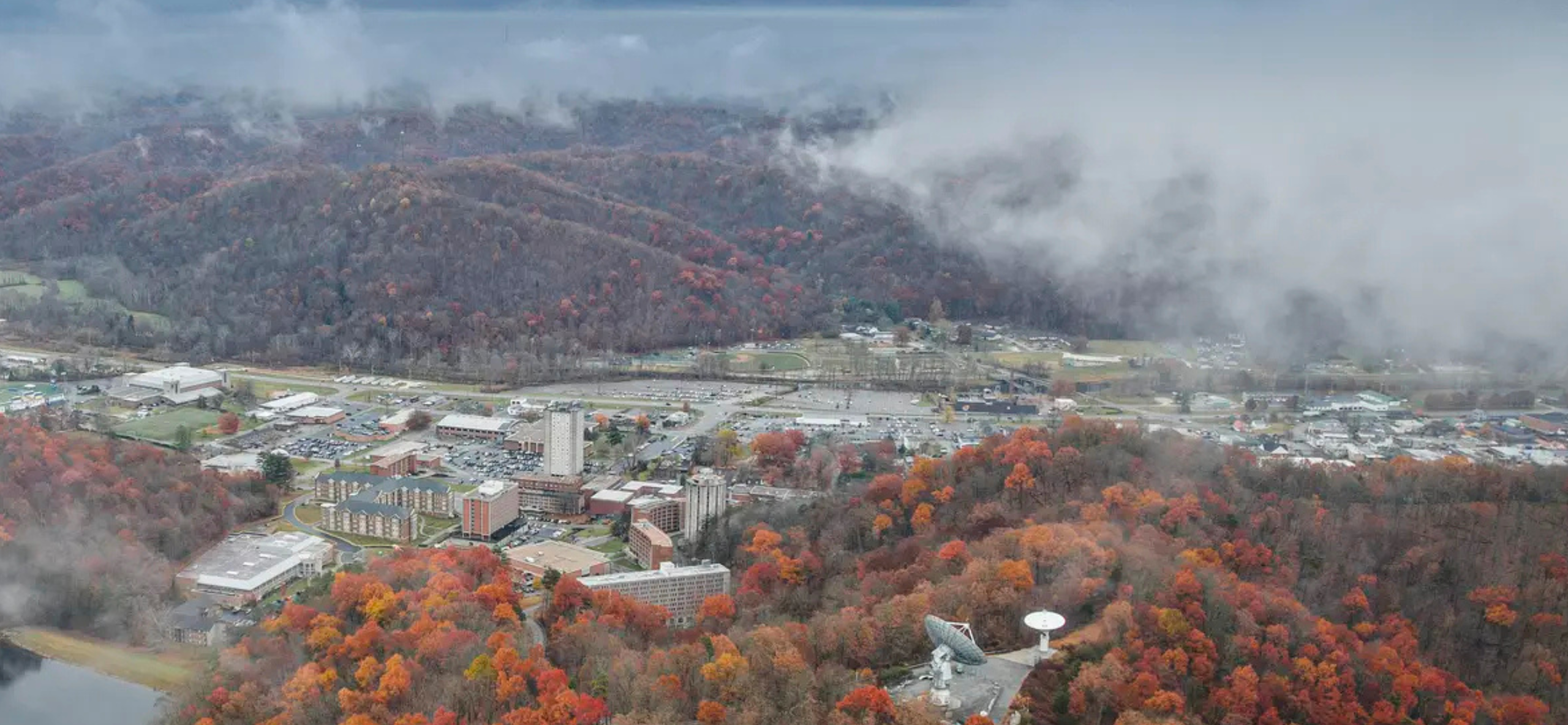 Aerial view of a campus in fall, surrounded by colorful trees and misty hills. Mid-rise buildings, parking lots, and satellite dishes are visible below.