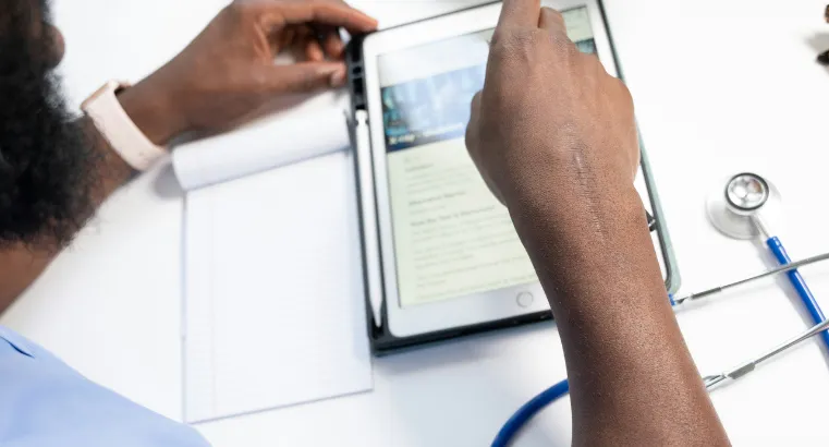 person in healthcare looking at tablet while notepad and stethoscope sit nearby.