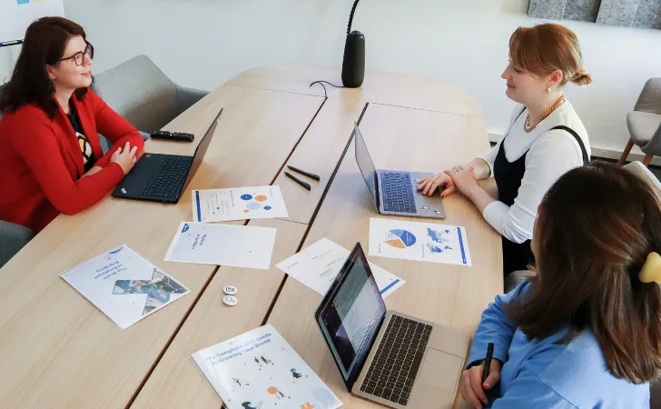Three people at a conference table with laptops and marketing documents.